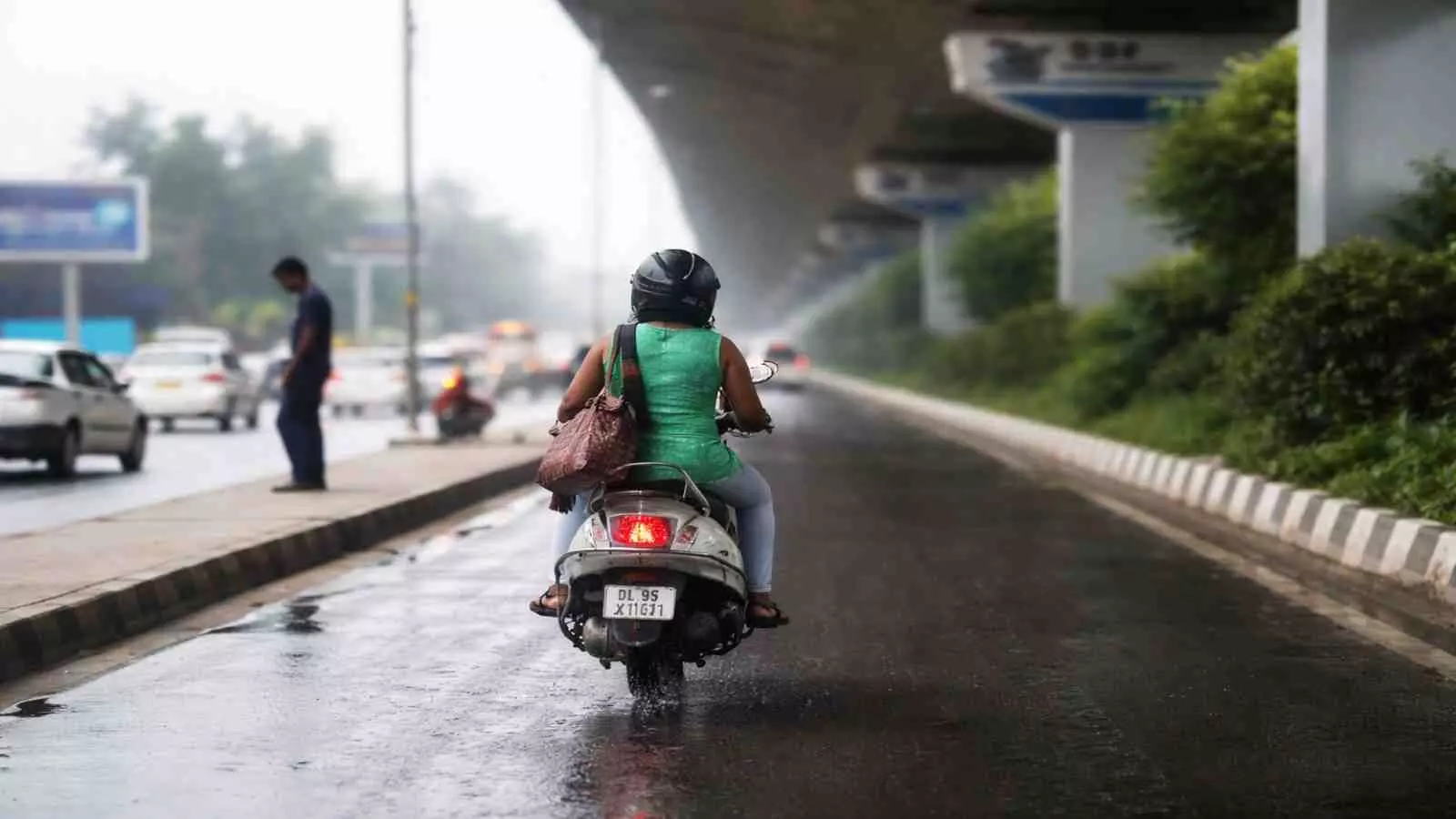 बारिश के बाद गीली सड़क पर स्कूटर चलाती महिला और पीछे से आती कार। बारिश के बाद गीली सड़क पर स्कूटर चलाती महिला और पीछे से आती कार।