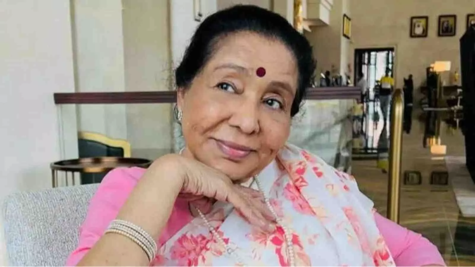 फूलों वाली सफेद साड़ी पहने मुस्कुराती हुई आशा भोसले कुर्सी पर बैठी हैं।। फूलों वाली सफेद साड़ी पहने मुस्कुराती हुई आशा भोसले कुर्सी पर बैठी हैं।।