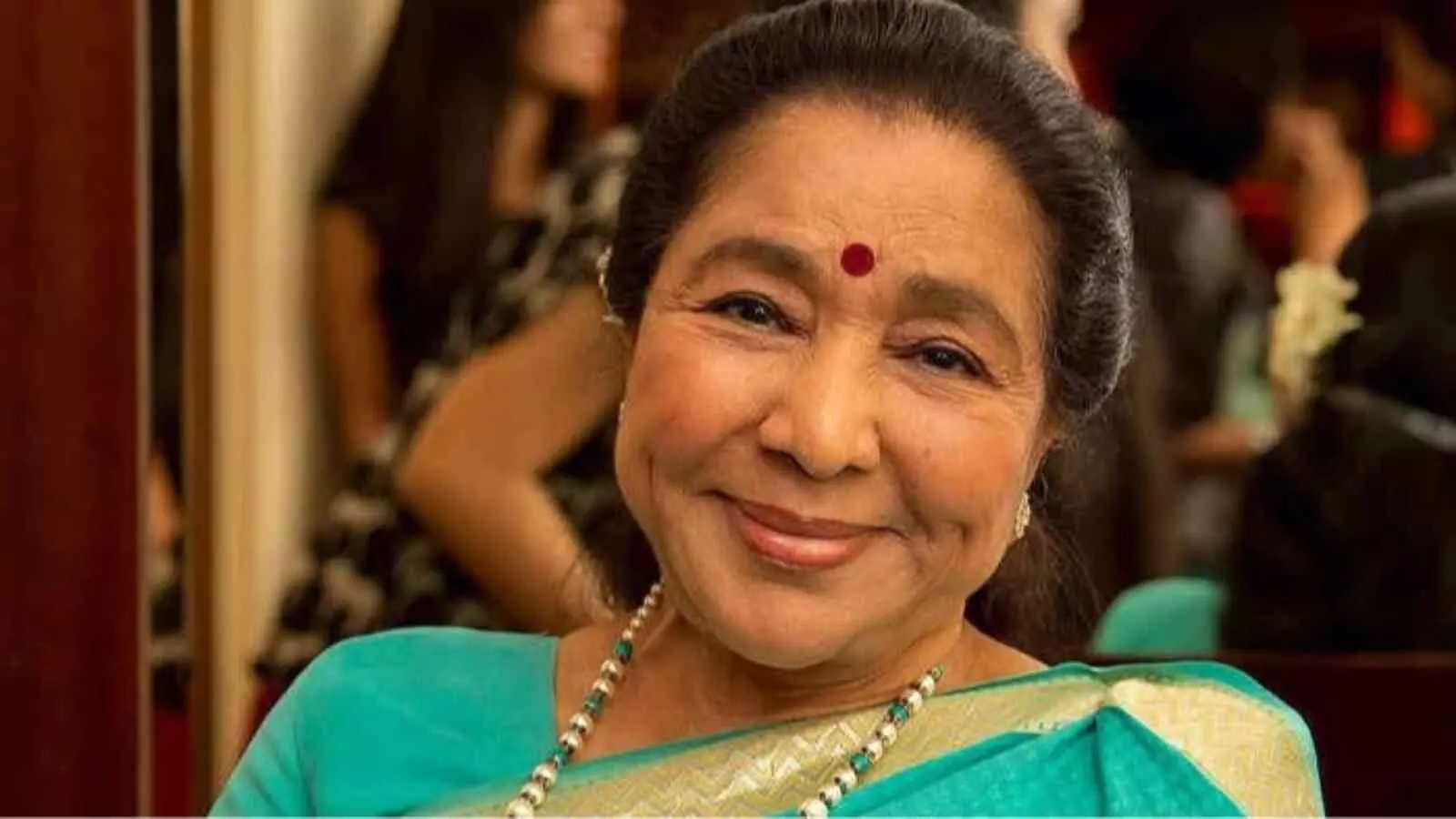 फिरोजी रंग की साड़ी और गले में मोतियों की माला पहने आशा भोसले मुस्कुरा रही हैं। फिरोजी रंग की साड़ी और गले में मोतियों की माला पहने आशा भोसले मुस्कुरा रही हैं।
