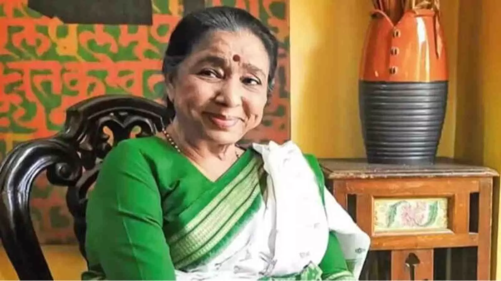 हरी और सफेद साड़ी पहने आशा भोसले काली नक्काशीदार कुर्सी पर बैठकर मुस्कुरा रही हैं। हरी और सफेद साड़ी पहने आशा भोसले काली नक्काशीदार कुर्सी पर बैठकर मुस्कुरा रही हैं।