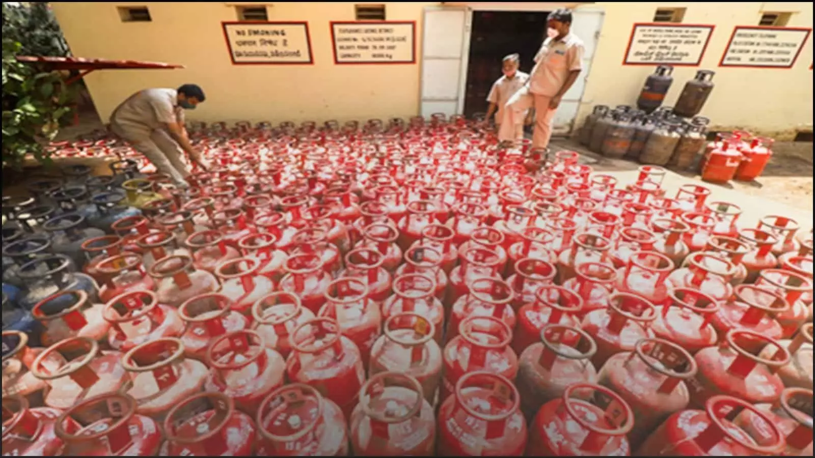 गैस गोदाम में रखे लाल रंग के अनेक घरेलू एलपीजी सिलेंडर और काम करते हुए कर्मचारी।
