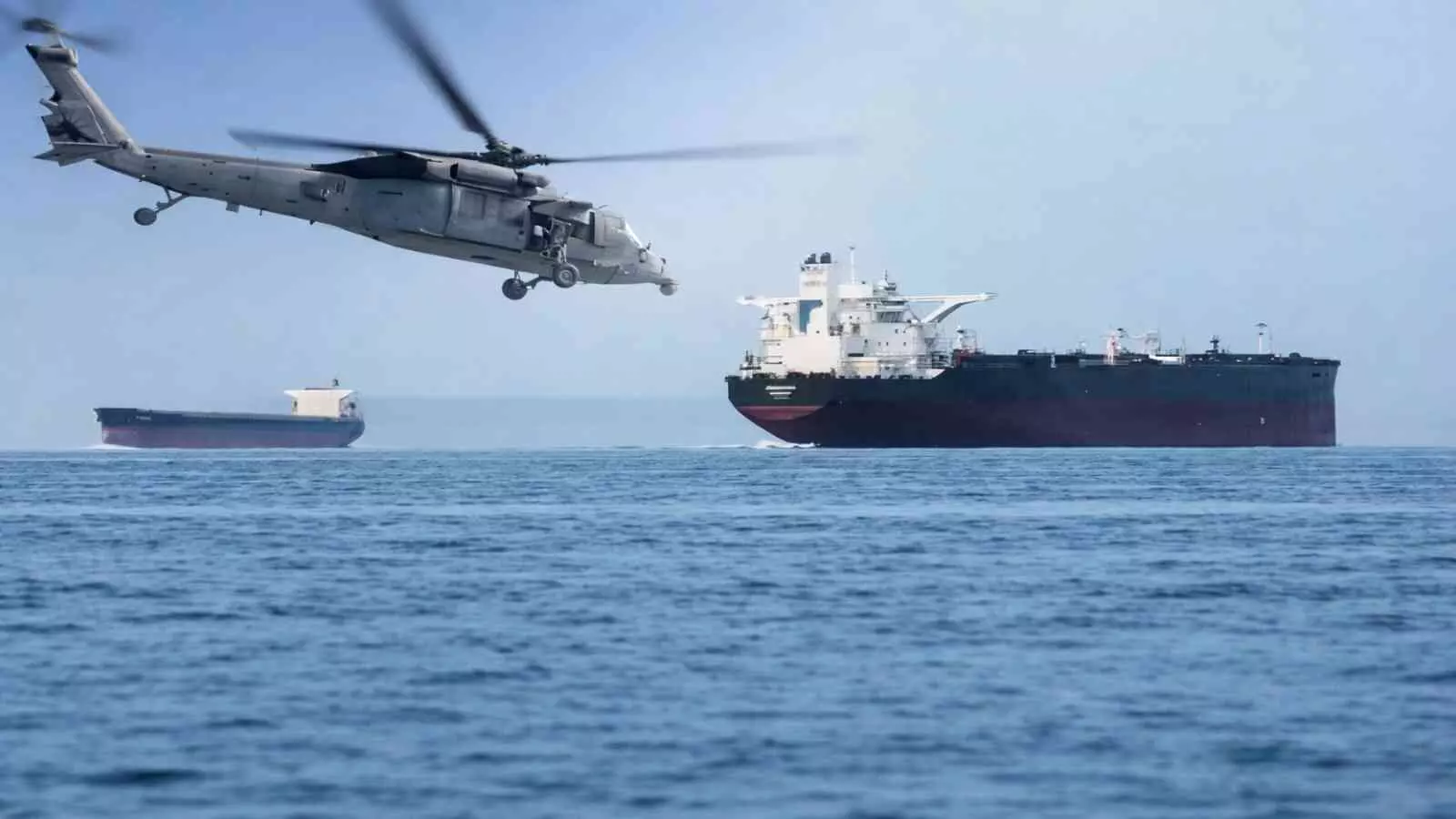 अरब सागर के ऊपर उड़ता सैन्य हेलीकॉप्टर और होर्मुज मार्ग पर खड़े बड़े मालवाहक जहाज।