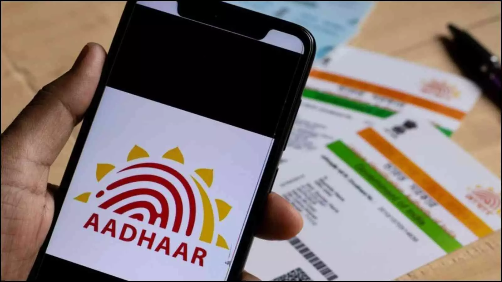 एक स्मार्टफोन स्क्रीन पर आधार का लोगो और पीछे भौतिक आधार कार्ड डिजिटल सत्यापन प्रक्रिया के संदर्भ में दिख रहे हैं।