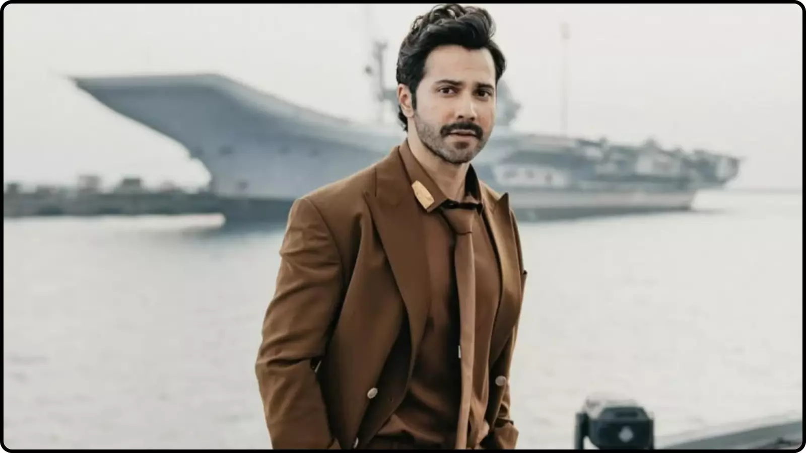 भूरे रंग के कोट और टाई पहने वरुण धवन एक विशाल समुद्री जहाज के सामने खड़े होकर देख रहे हैं।