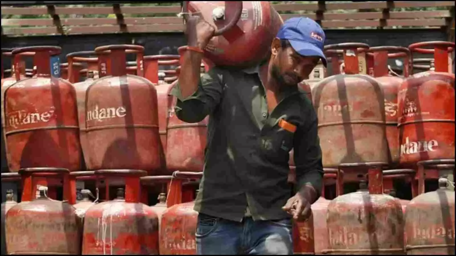 लाल रंग के इंडेन गैस सिलेंडरों के ढेर के सामने एक व्यक्ति कंधे पर सिलेंडर रखकर ले जा रहा है। लाल रंग के इंडेन गैस सिलेंडरों के ढेर के सामने एक व्यक्ति कंधे पर सिलेंडर रखकर ले जा रहा है।