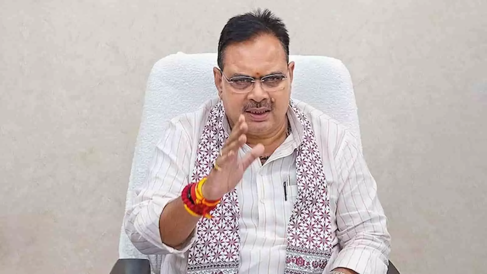 सफेद पृष्ठभूमि में कुर्सी पर बैठे राजस्थान के मुख्यमंत्री भजनलाल शर्मा ओसियां में किसान निधि जारी करते हुए।