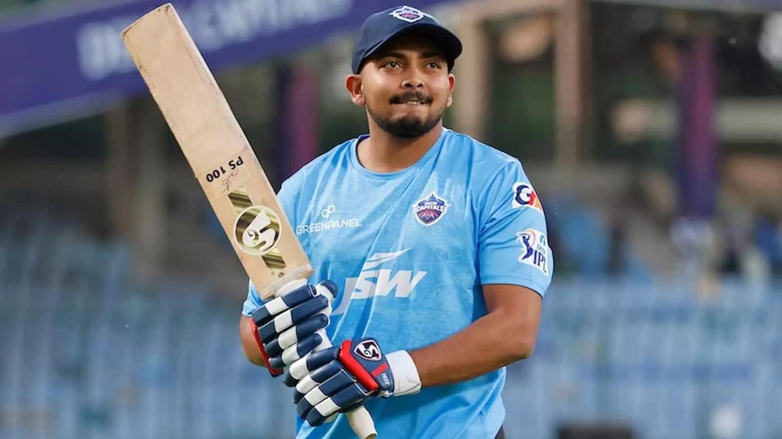 क्रिकेट के मैदान पर नीली जर्सी और हेलमेट पहने हुए बल्लेबाज पृथ्वी शॉ हाथ में बल्ला लिए खड़े हैं। क्रिकेट के मैदान पर नीली जर्सी और हेलमेट पहने हुए बल्लेबाज पृथ्वी शॉ हाथ में बल्ला लिए खड़े हैं।
