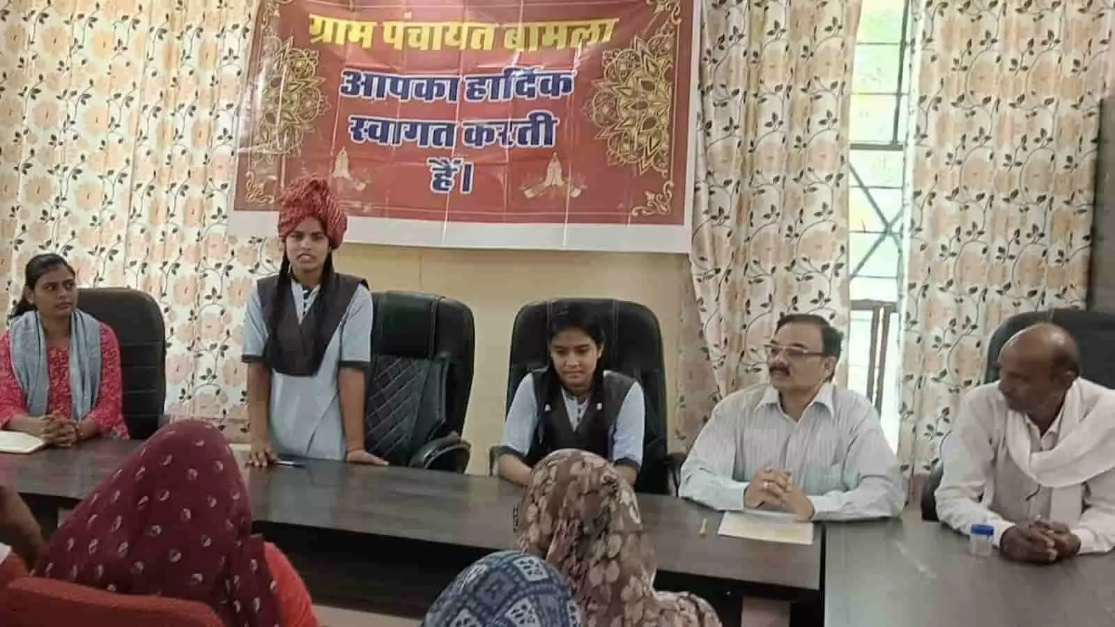 बामला ग्राम पंचायत में टेबल के पास बैठे अधिकारी और खड़े होकर बोलती छात्रा। बामला ग्राम पंचायत में टेबल के पास बैठे अधिकारी और खड़े होकर बोलती छात्रा।