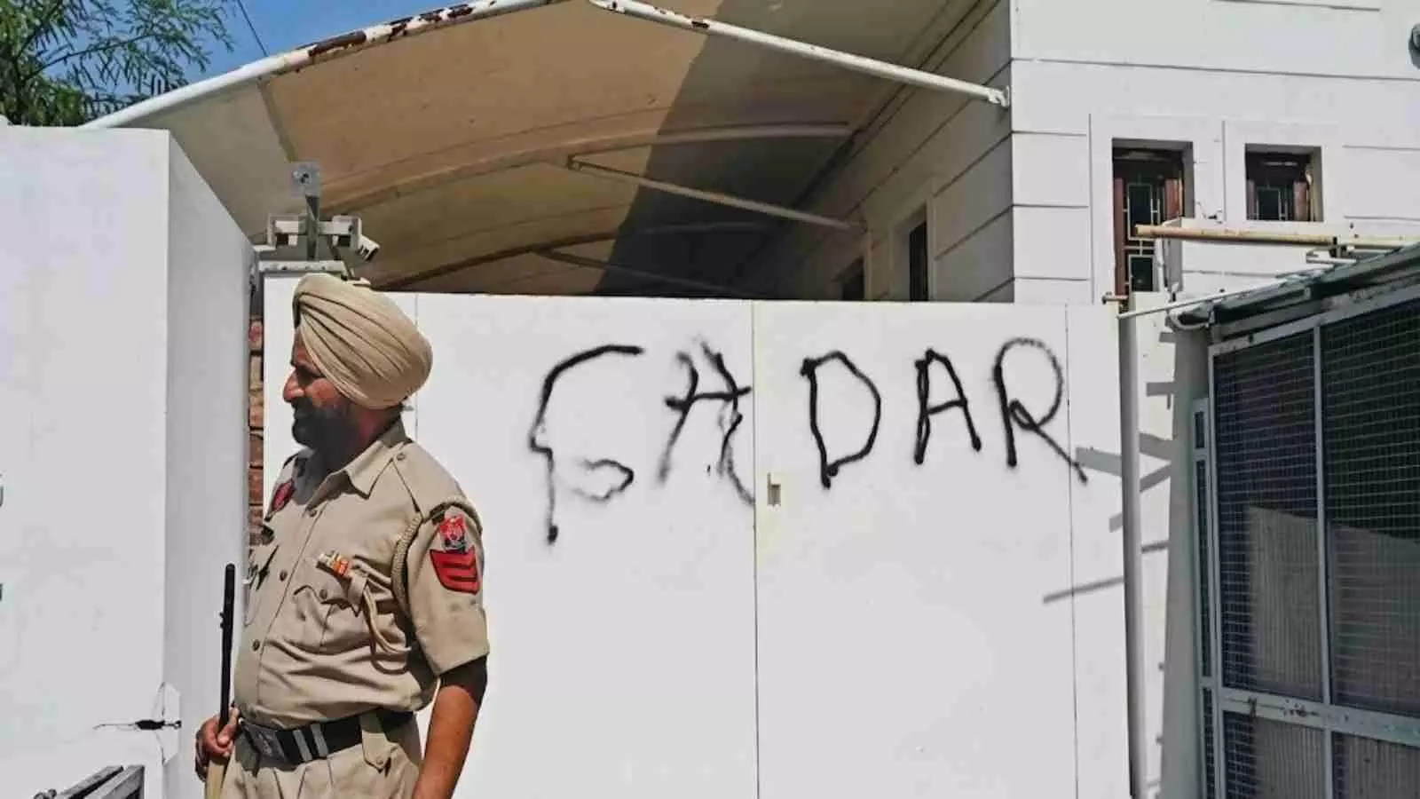 सफेद दीवार पर काले रंग से गद्दार लिखा हुआ है और पास में पंजाब पुलिस का जवान खड़ा है। सफेद दीवार पर काले रंग से गद्दार लिखा हुआ है और पास में पंजाब पुलिस का जवान खड़ा है।