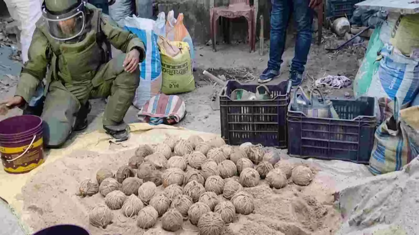 जमीन पर रखे हुए गोल आकार के बम और पास में बम निरोधक सूट पहने सुरक्षाकर्मी जांच करते दिख रहे हैं। जमीन पर रखे हुए गोल आकार के बम और पास में बम निरोधक सूट पहने सुरक्षाकर्मी जांच करते दिख रहे हैं।