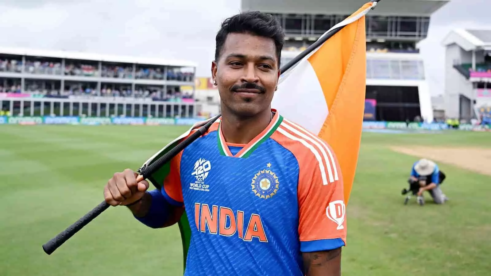 टी20 विश्व कप जर्सी में हार्दिक पांड्या कंधे पर भारतीय झंडा रखे हुए क्रिकेट मैदान पर खड़े हैं।