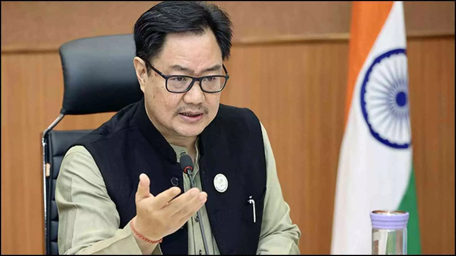 एक मेज पर रखे माइक के सामने चश्मा पहने किरेन रिजिजू हाथ के इशारे से अपनी बात समझाते हुए।