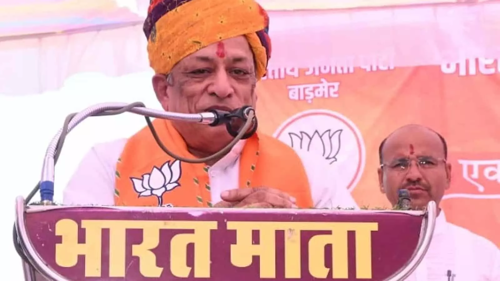 केसरिया साफा पहने कैबिनेट मंत्री जोराराम कुमावत मंच पर माइक के सामने भाषण देते हुए।