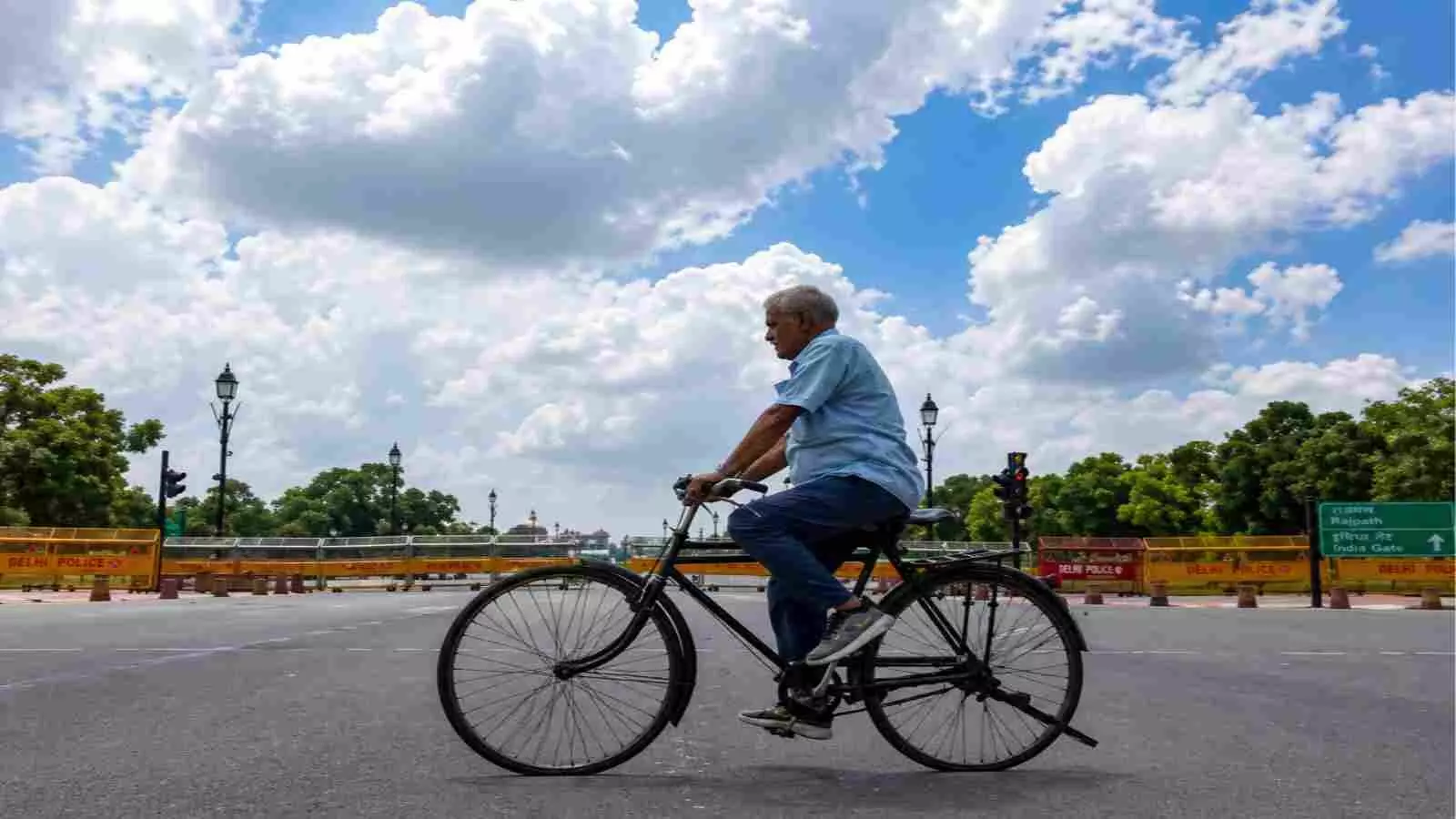 नीले आसमान और घने सफेद बादलों के नीचे सड़क पर साइकिल चलाता हुआ एक बुजुर्ग व्यक्ति।