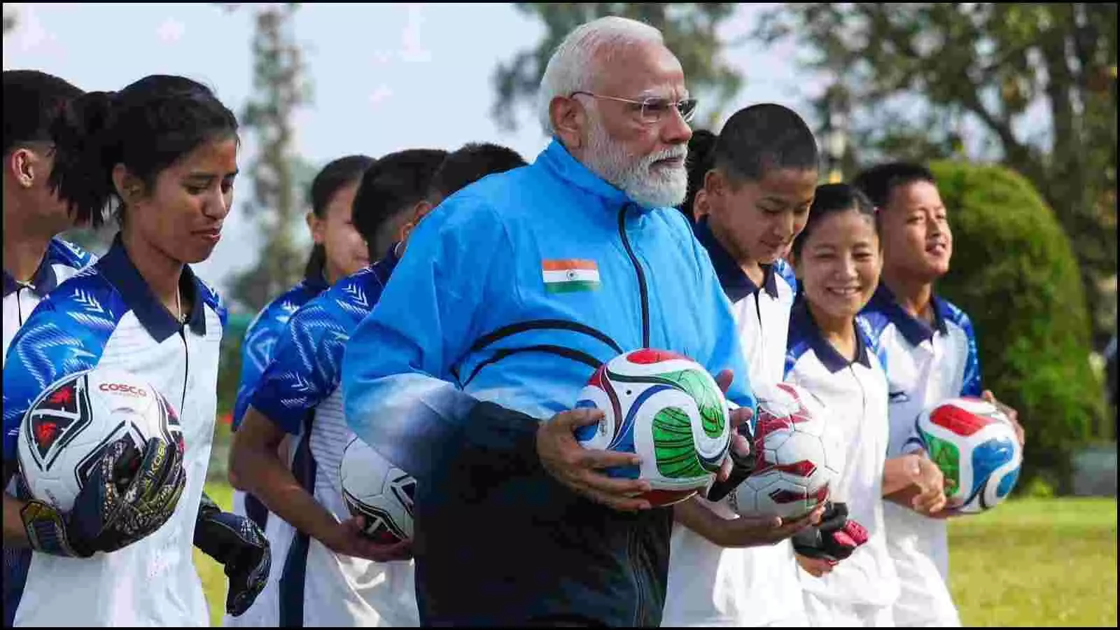 नीले ट्रैक सूट में प्रधानमंत्री नरेंद्र मोदी गंगटोक के मैदान पर बच्चों के साथ फुटबॉल पकड़े हुए।