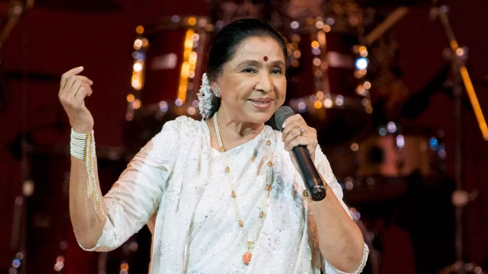हाथ में माइक थामे सफेद साड़ी पहने आशा भोसले स्टेज पर लाइव संगीत कार्यक्रम में गा रही हैं।