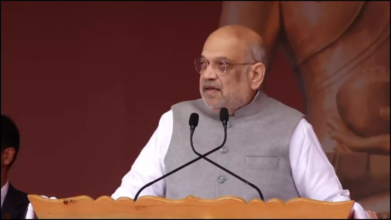 पोडियम के पीछे खड़े होकर चश्मा पहने अमित शाह लेह में बुद्ध प्रदर्शनी के दौरान भाषण दे रहे हैं।