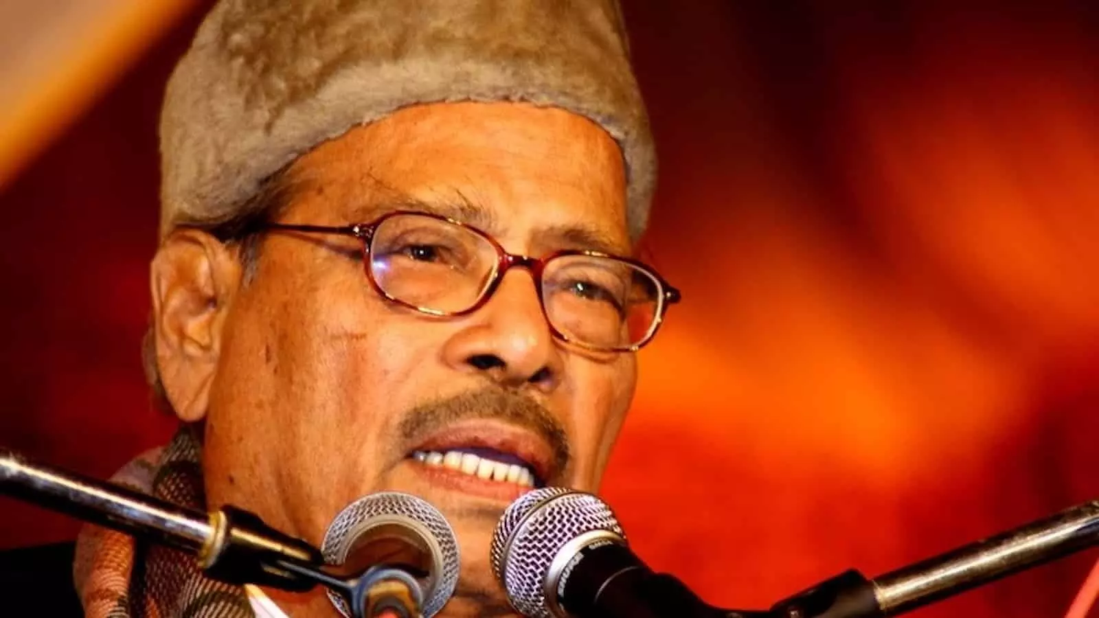 चश्मा और टोपी पहने गायक मन्ना डे माइक्रोफोन के सामने मंच पर गाना गा रहे हैं।