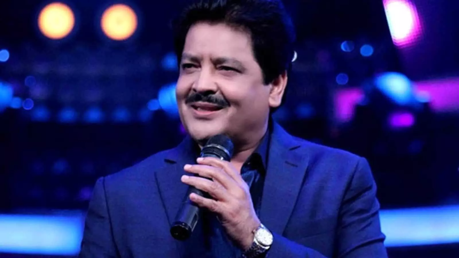 हाथ में माइक लिए मुस्कुराते हुए गायक उदित नारायण एक स्टेज शो के दौरान गाना गा रहे हैं।