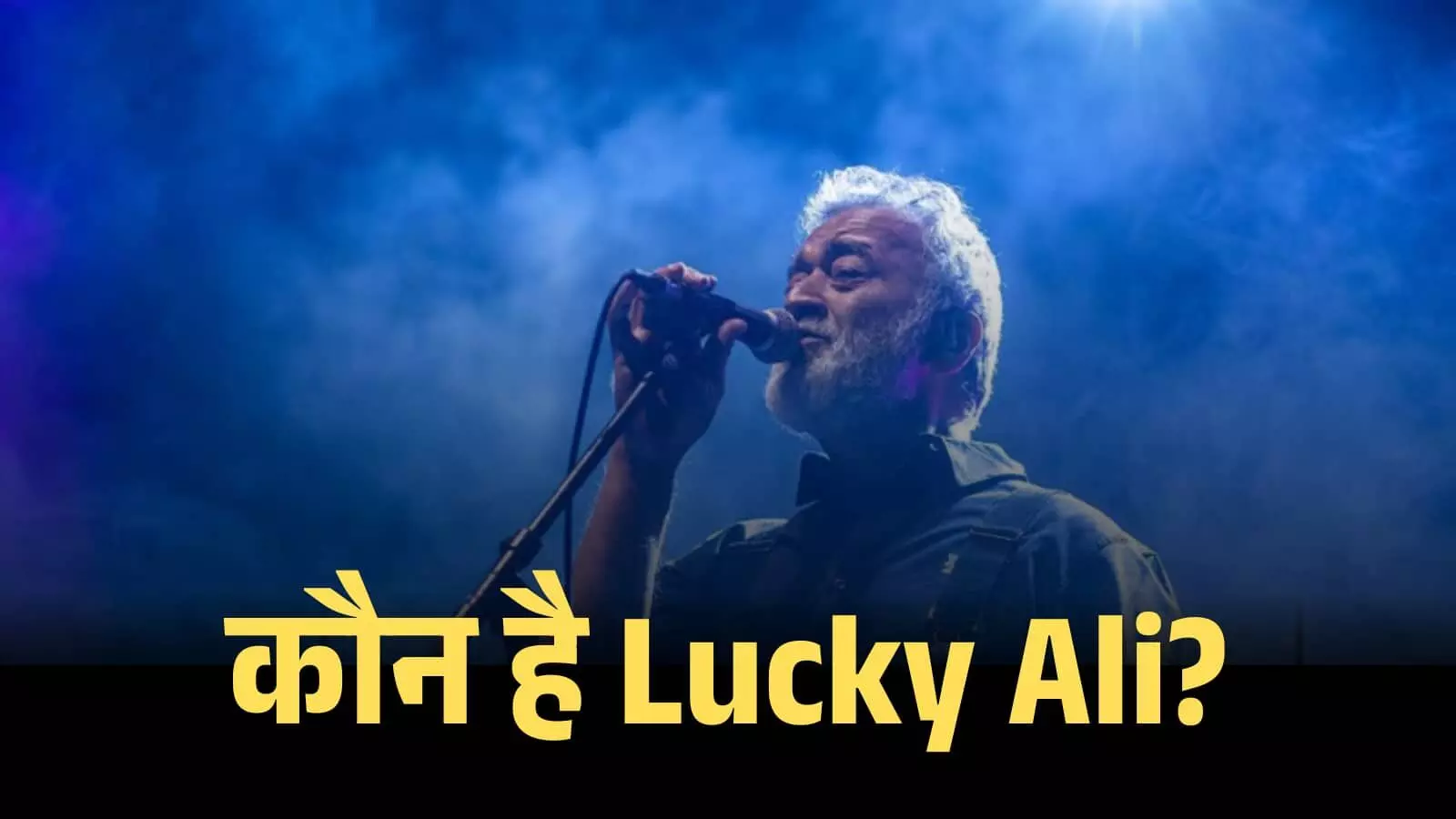 एक संगीत समारोह के दौरान नीली रोशनी में मंच पर माइक थामे गाते हुए गायक लकी अली। एक संगीत समारोह के दौरान नीली रोशनी में मंच पर माइक थामे गाते हुए गायक लकी अली।