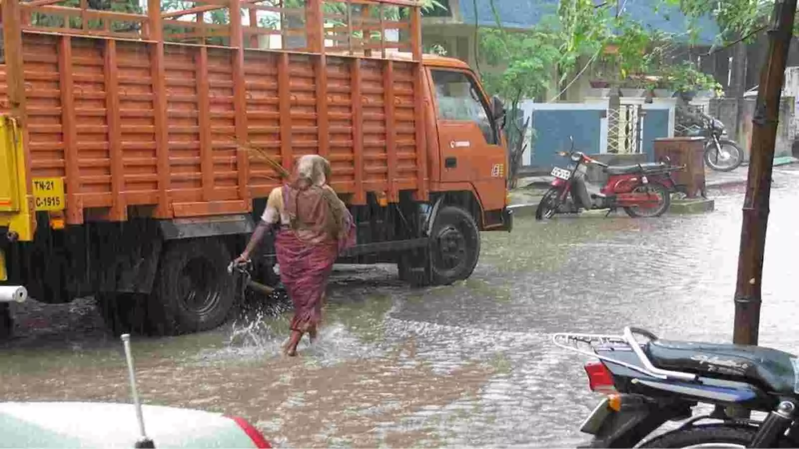 भारी बारिश के दौरान जलभराव वाली सड़क पर एक महिला पैदल चलती हुई और पास में खड़ा नारंगी रंग का ट्रक। भारी बारिश के दौरान जलभराव वाली सड़क पर एक महिला पैदल चलती हुई और पास में खड़ा नारंगी रंग का ट्रक।