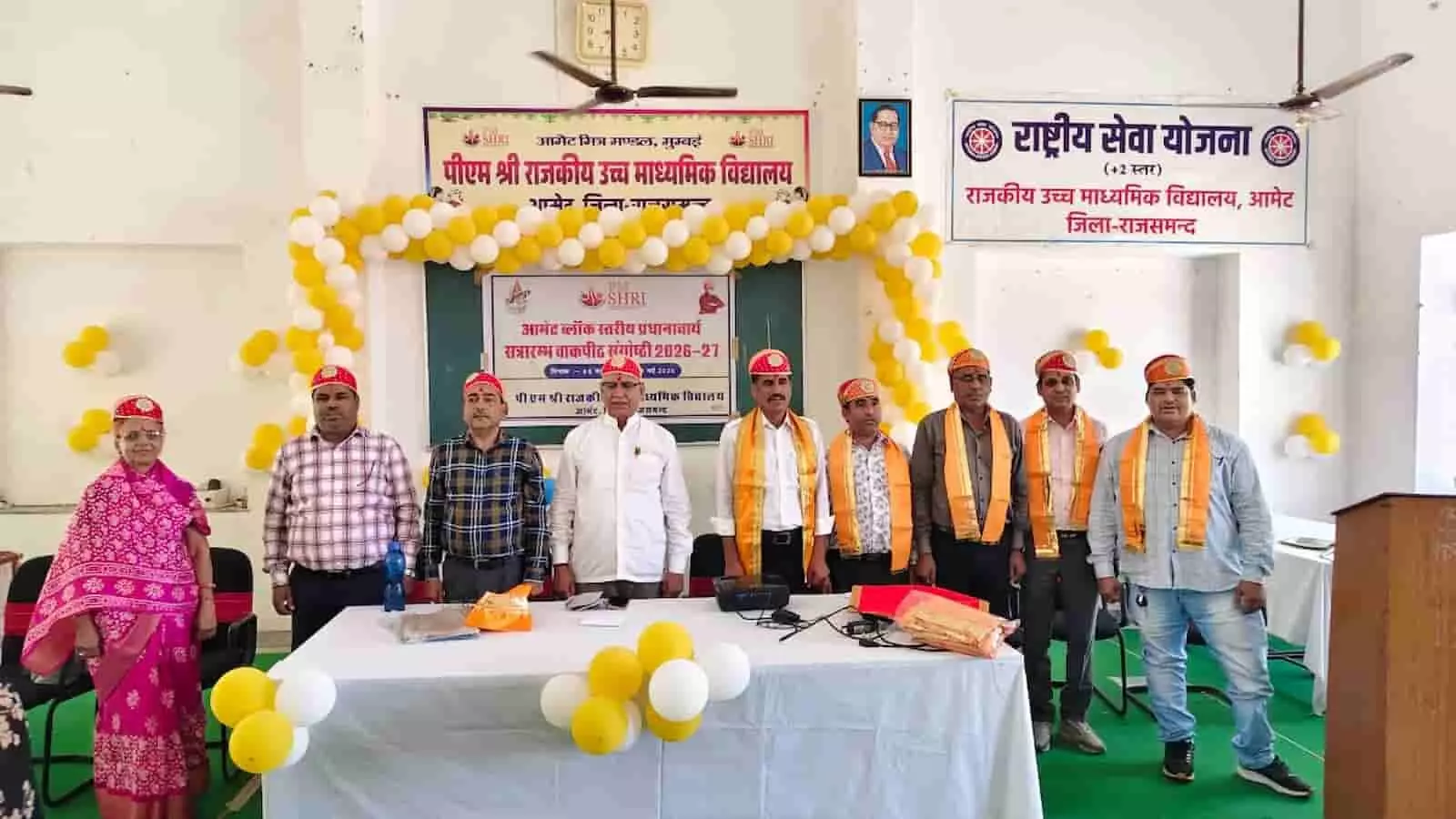 आमेट के विद्यालय सभागार में मंच पर खड़े माला और साफा पहने हुए प्रधानाचार्य और शिक्षा अधिकारी।