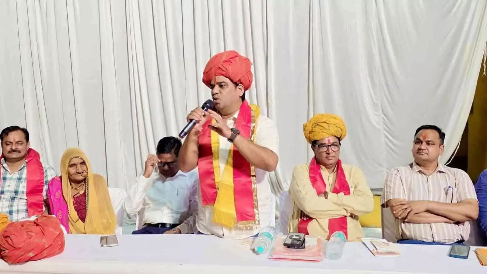 रात्रि चौपाल के दौरान मंच पर माइक से ग्रामीणों को संबोधित करते हुए भाजपा नेता और अन्य अधिकारी।