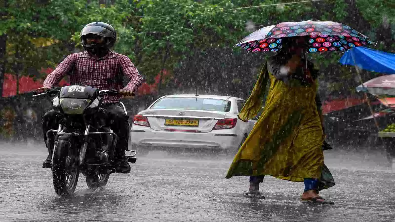 बारिश के दौरान सड़क पर छतरी लेकर चलती महिला और रेनकोट पहने बाइक सवार व्यक्ति। बारिश के दौरान सड़क पर छतरी लेकर चलती महिला और रेनकोट पहने बाइक सवार व्यक्ति।