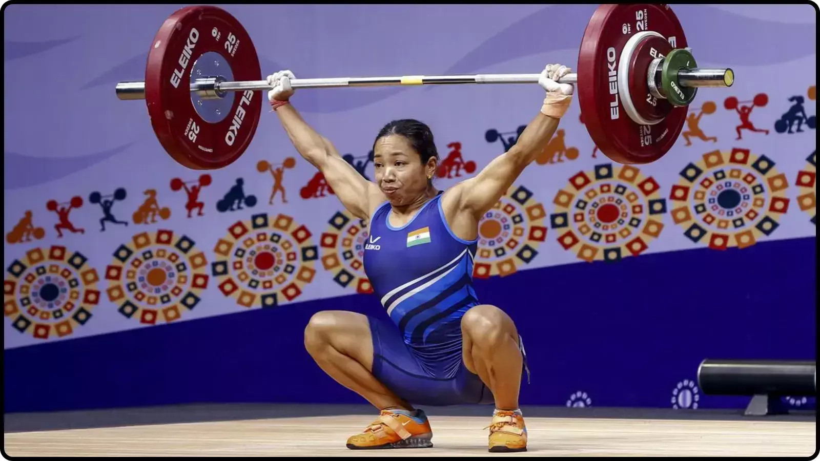 नीले रंग की जर्सी पहनी एक महिला खिलाड़ी वेटलिफ्टिंग एरिना में भारी बारबेल उठाए हुए। नीले रंग की जर्सी पहनी एक महिला खिलाड़ी वेटलिफ्टिंग एरिना में भारी बारबेल उठाए हुए।