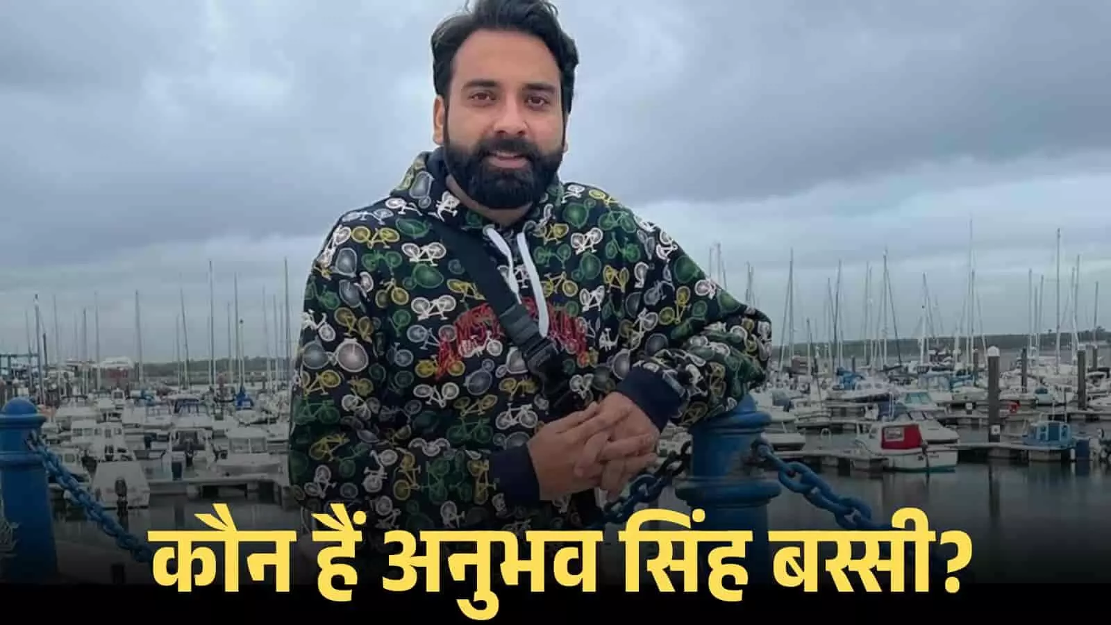 नीली और काली हुडी पहने अनुभव सिंह बस्सी समुद्र किनारे नावों के सामने खड़े होकर मुस्कुरा रहे हैं।