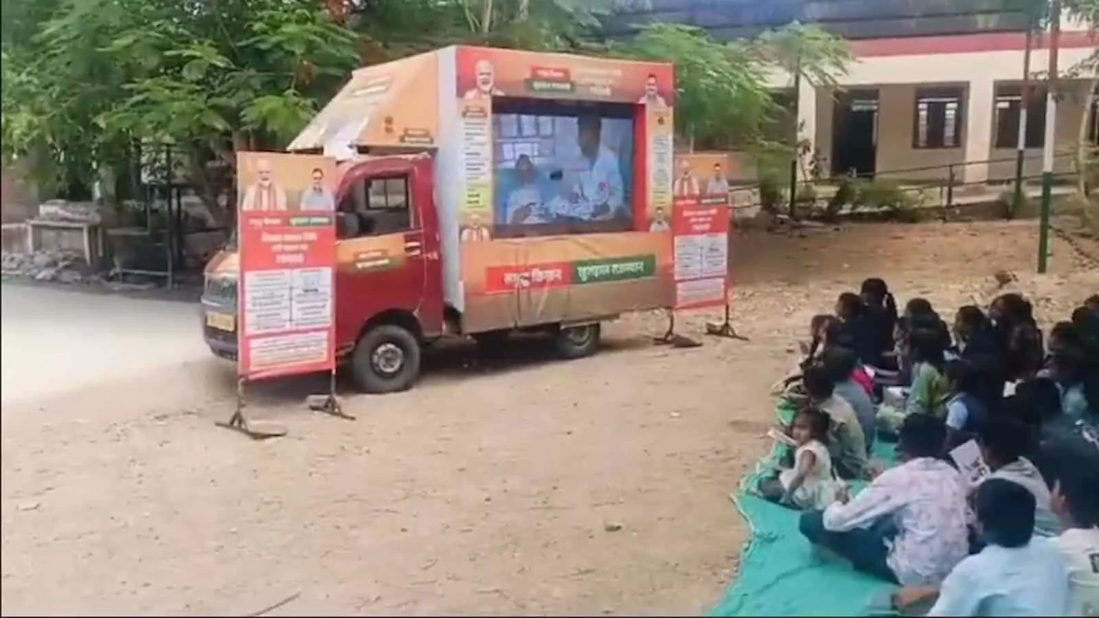 अर्जुनगढ़ के स्कूल परिसर में विकास रथ यात्रा की एलईडी वैन के सामने जमीन पर बैठे स्कूली बच्चे।