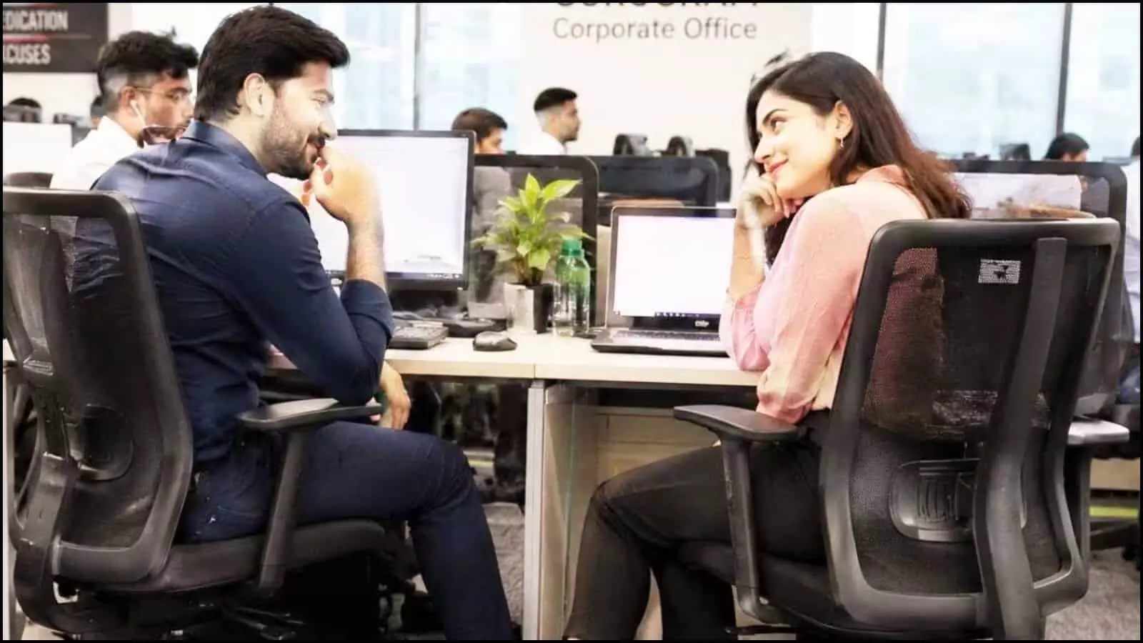 ऑफिस डेस्क पर कंप्यूटर के सामने बैठकर मुस्कुराते हुए एक-दूसरे को देखते सहकर्मी वर्कप्लेस रोमांस के संदर्भ में।