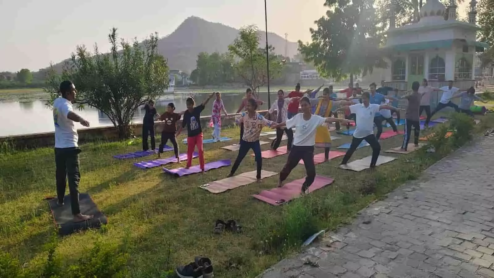 सलूंबर में तालाब के किनारे हरी घास पर योग मैट बिछाकर सामूहिक योगाभ्यास करते हुए लोग और योग प्रशिक्षक।