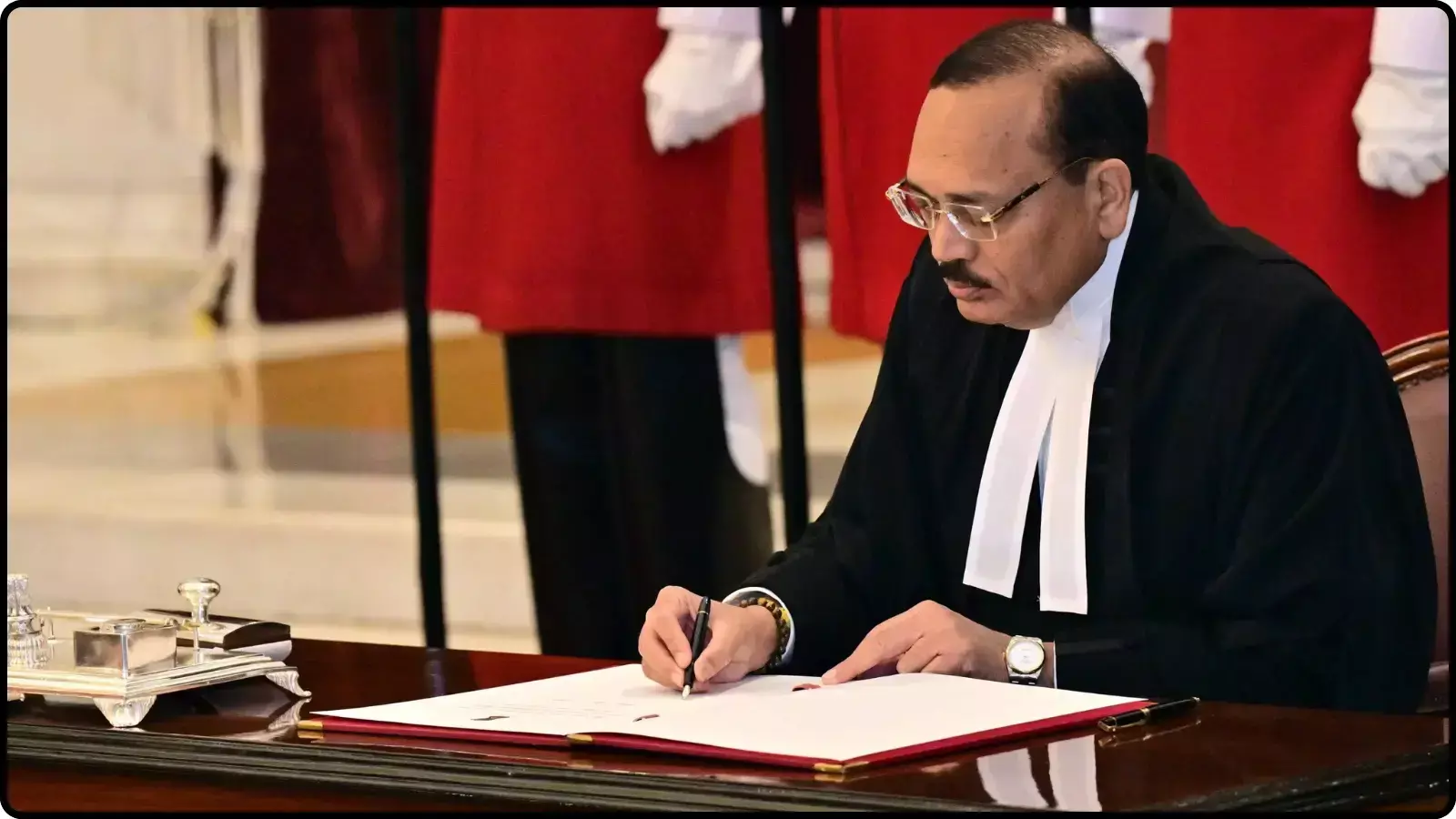 मेज पर रखे सफेद कागज पर पेन से हस्ताक्षर करते हुए काले गाउन पहने एक न्यायाधीश। मेज पर रखे सफेद कागज पर पेन से हस्ताक्षर करते हुए काले गाउन पहने एक न्यायाधीश।