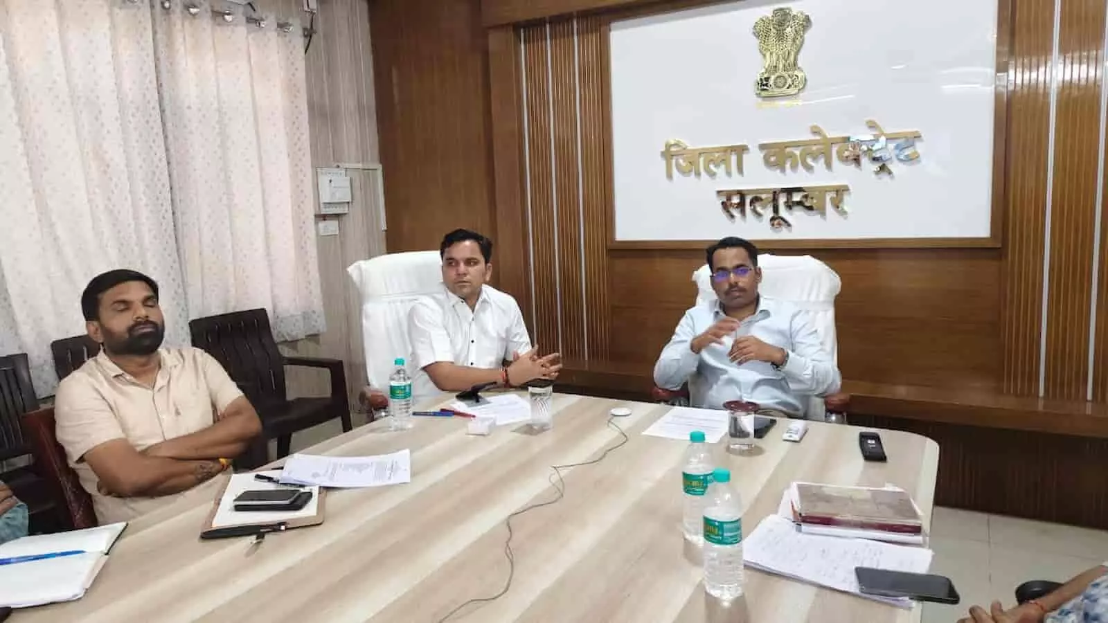 जिला कलेक्ट्रेेट सलूम्बर के बैठक कक्ष में बैठे अधिकारी और दीवार पर लगा राज्य प्रतीक चिह्न।