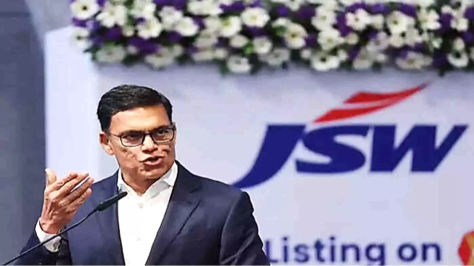 एक औपचारिक कोट पहने हुए सज्जन जिंदल जेएसडब्ल्यू लोगो के सामने माइक पर बोलते हुए। एक औपचारिक कोट पहने हुए सज्जन जिंदल जेएसडब्ल्यू लोगो के सामने माइक पर बोलते हुए।