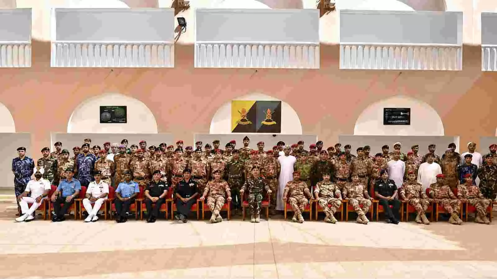 सैन्य वर्दी पहने हुए भारत और ओमान के कई सैनिक एक भवन के सामने कुर्सियों पर बैठे हैं।