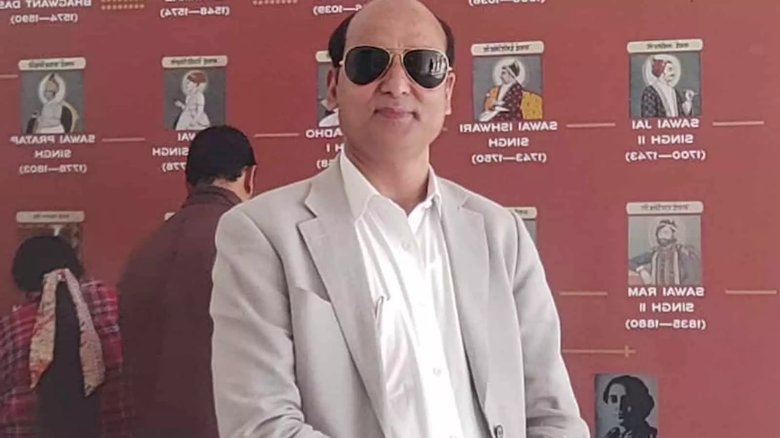 चश्मा और कोट पहने हुए डॉक्टर शमशाद अली एक संग्रहालय या गैलरी में प्रदर्शित वंशावली चार्ट बोर्ड के सामने खड़े हैं।