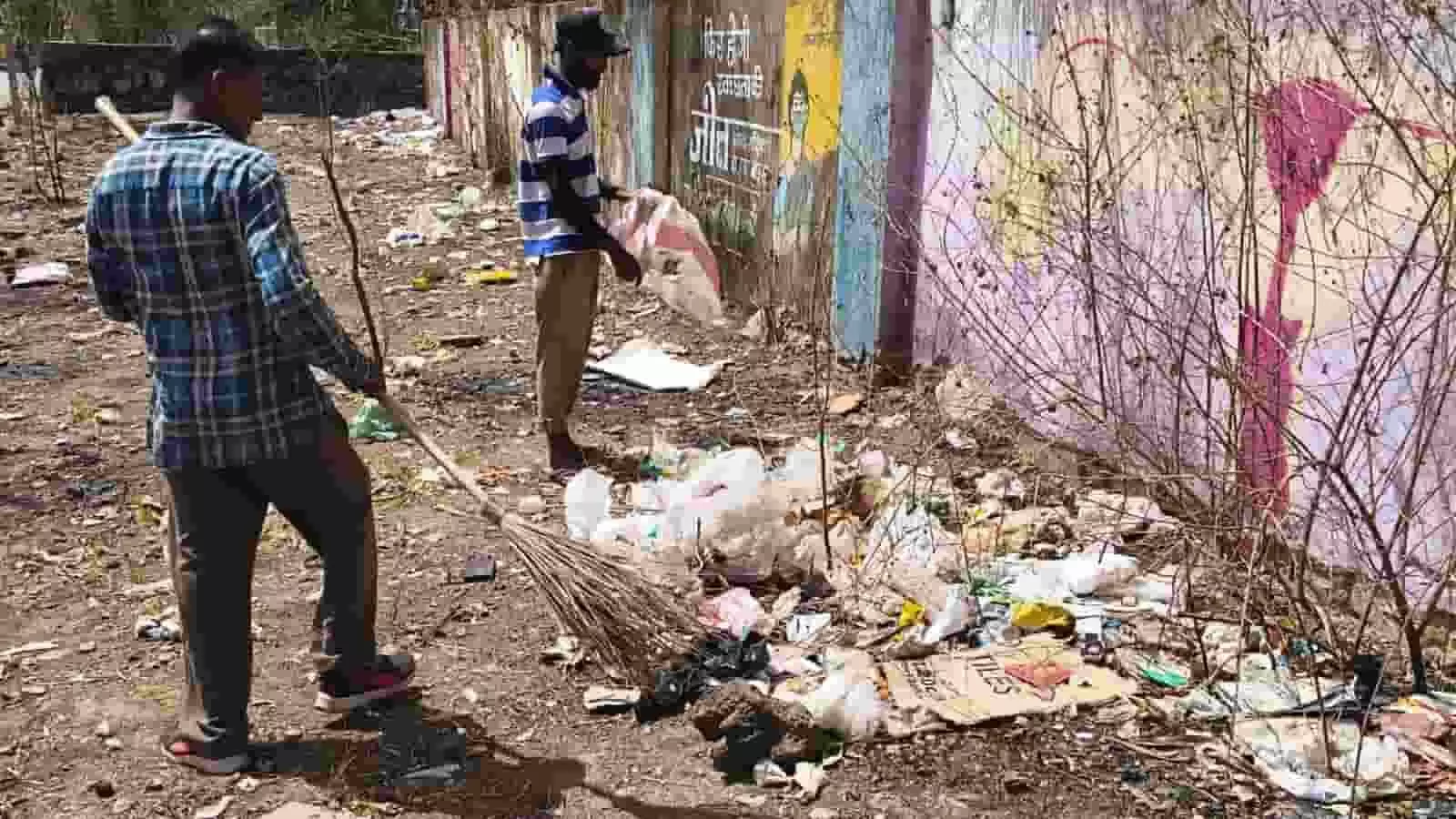 खुले मैदान में झाड़ू और बोरी लेकर कचरा साफ करते हुए दो व्यक्ति उदयपुर में स्वच्छता अभियान के दौरान दिख रहे हैं।