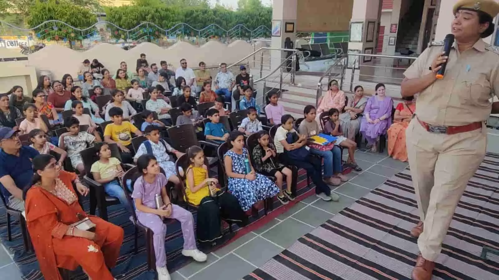 अभिरुचि शिविर में माइक हाथ में लेकर बच्चों को सड़क सुरक्षा नियम समझातीं पुलिस यूनिफॉर्म में खड़ी इंस्पेक्टर टीना सोलंकी और कुर्सियों पर बैठे बच्चे।