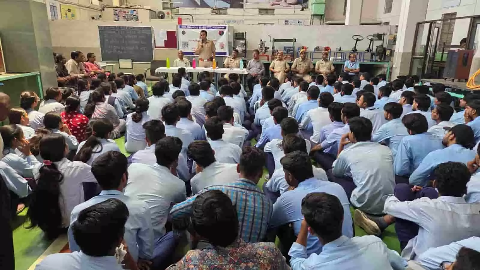 राजकीय औद्योगिक प्रशिक्षण संस्थान भीलवाड़ा परिसर में जमीन पर बैठे विद्यार्थियों को संबोधित करते हुए मंच पर मौजूद वर्दीधारी पुलिस अधिकारी।
