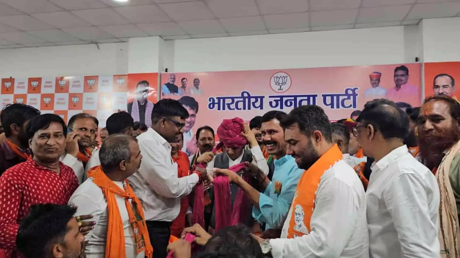 भाजपा कार्यालय में कार्यकर्ताओं से घिरे नेता कैलाश चौधरी सिर पर गुलाबी पगड़ी बांधते हुए भीलवाड़ा स्वागत समारोह में उपस्थित हैं।