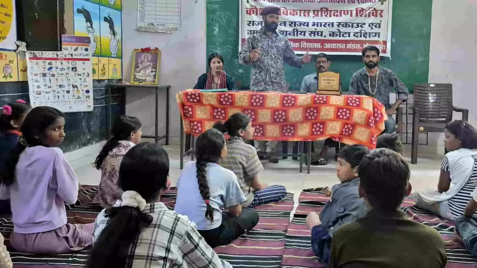 कक्षा में दरी पर बैठे विद्यार्थियों को सामने टेबल के पास खड़े होकर संबोधित करते वक्ता। कक्षा में दरी पर बैठे विद्यार्थियों को सामने टेबल के पास खड़े होकर संबोधित करते वक्ता।