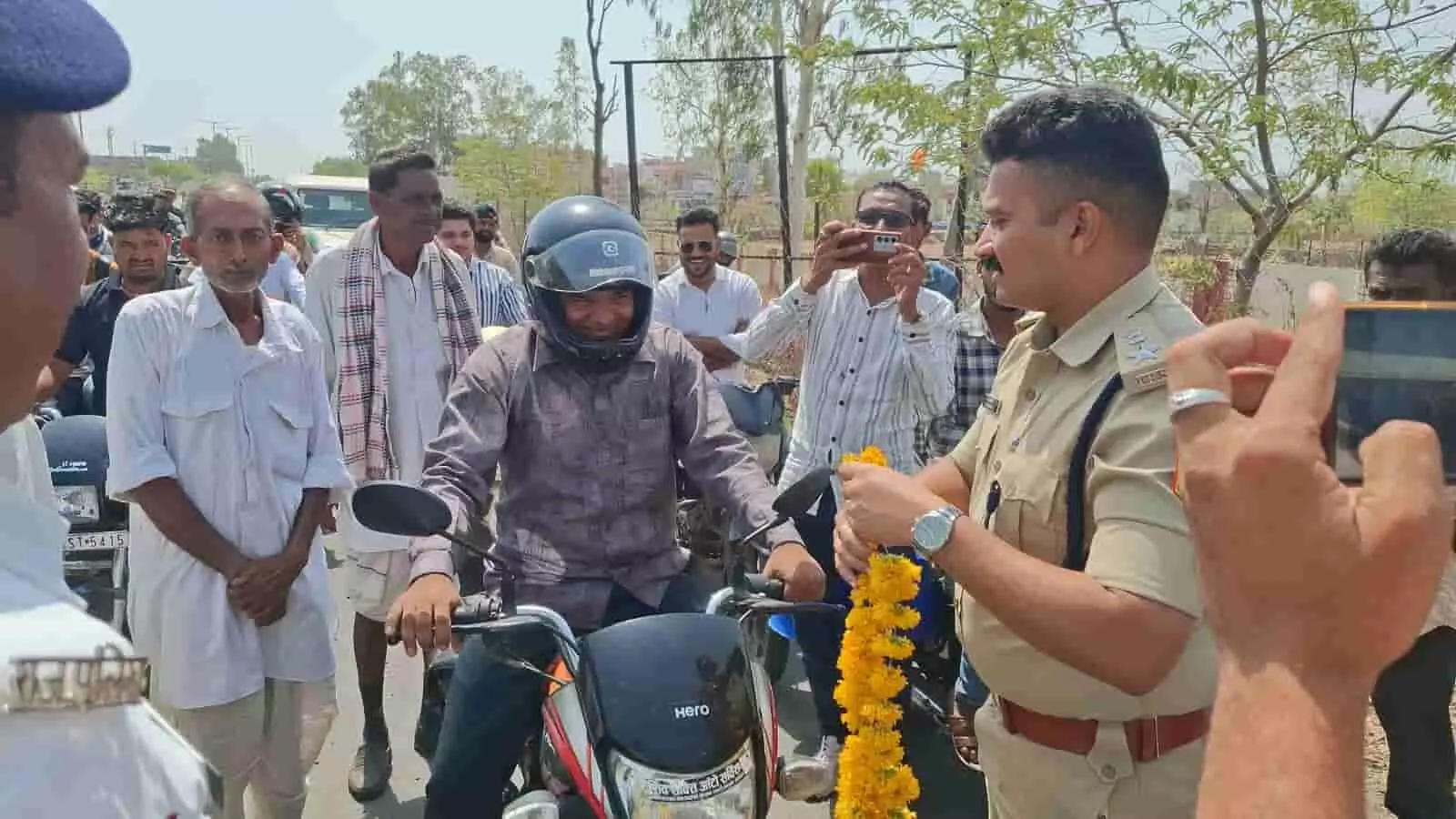 सड़क पर मोटरसाइकिल सवार हेलमेट पहने व्यक्ति को माला पहनाते पुलिस अधिकारी और खड़े लोग। सड़क पर मोटरसाइकिल सवार हेलमेट पहने व्यक्ति को माला पहनाते पुलिस अधिकारी और खड़े लोग।