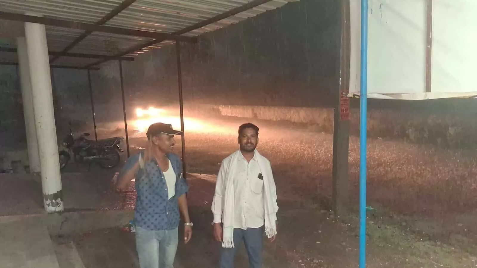 मूसलाधार बारिश के दौरान टिन शेड के नीचे खड़े दो पुरुष लसाडिया में मौसम परिवर्तन के संदर्भ में दिख रहे हैं। मूसलाधार बारिश के दौरान टिन शेड के नीचे खड़े दो पुरुष लसाडिया में मौसम परिवर्तन के संदर्भ में दिख रहे हैं।