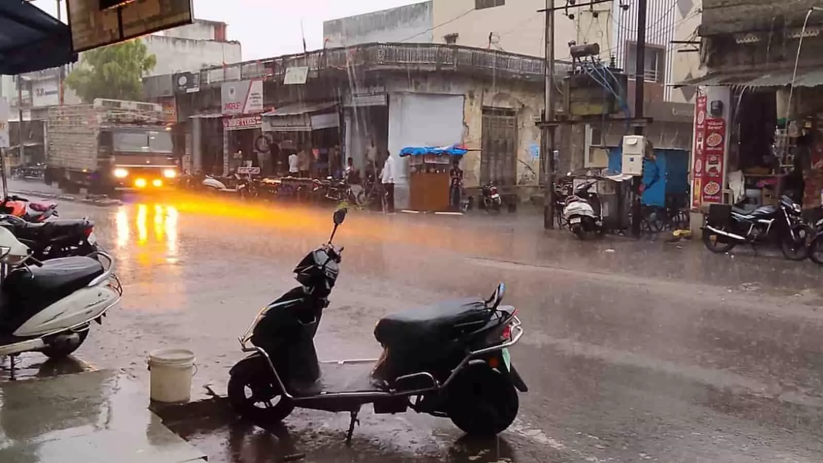 बारिश से भीगती फतहनगर की सड़क पर खड़े दोपहिया वाहन और गुजरते ट्रक का दृश्य।