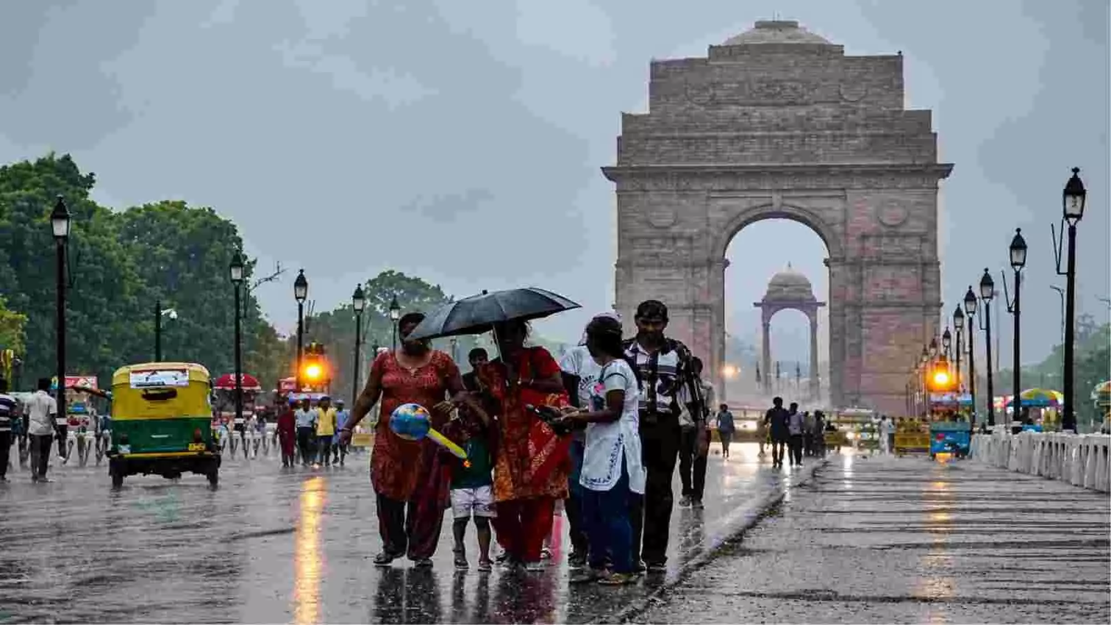 इंडिया गेट के सामने बारिश और आंधी के बीच छतरी पकड़कर सड़क पर चलते हुए लोग।