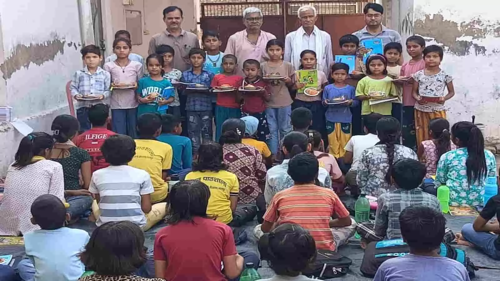 निशुल्क बाल पाठशाला में खड़े बच्चे हाथों में कॉपियां और नाश्ता लेकर शिक्षकों के साथ भरतपुर में समूह चित्र खिंचवा रहे हैं।