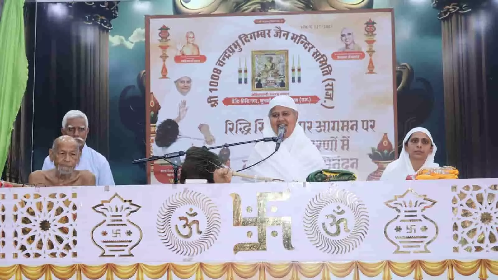 मंच पर माइक के सामने बैठकर प्रवचन देतीं जैन आर्यिका स्वस्ति भूषण माताजी और पास बैठे अन्य जैन साधक। मंच पर माइक के सामने बैठकर प्रवचन देतीं जैन आर्यिका स्वस्ति भूषण माताजी और पास बैठे अन्य जैन साधक।