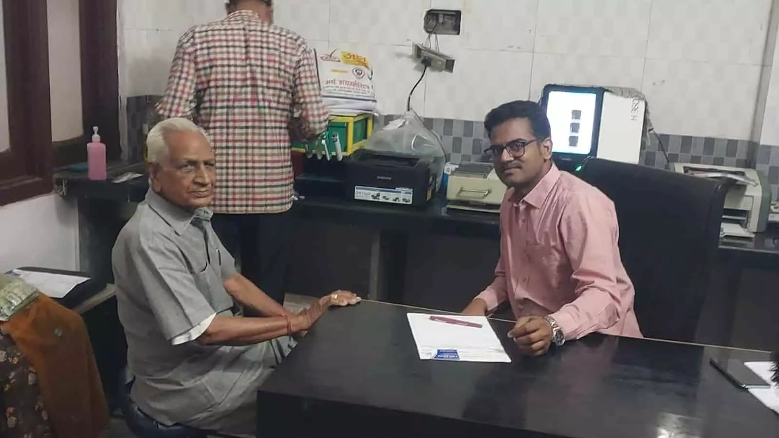 डायग्नोस्टिक सेंटर के केबिन में मेज पर बैठे डॉक्टर और परामर्श लेता बुजुर्ग मरीज।